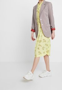 Persona che indossa un abito midi floreale giallo, un blazer a scacchi chiari con bordi rossi e sneaker bianche chunky, in piedi su uno sfondo bianco.