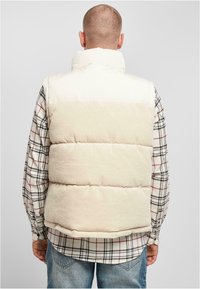 Uppblåsbar väst i krämfärgad och beige kordflors med hög krage, bärs över en rutigt skjorta. Har en vadderad design och strukturerad tyg.