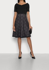 Černé šaty s krátkými rukávy, výstřihem do tvaru V a krajkovou sukní s květinovým vzorem. Doplněné černými lodičkami a psaníčkem.