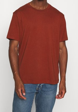 Mand iført en ensfarvet rustfarvet kortærmet T-shirt og blå denimjeans foran en hvid baggrund.