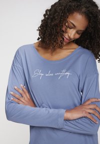 Long-sleeve, loose-fitting top in soft periwinkle blue fabric. Features a wide neckline and white handwritten text: "Sleep solves everything."