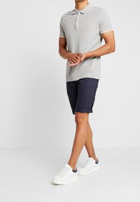 Uomo in piedi con le mani incrociate, che indossa una polo grigia chiara, pantaloncini scuri con motivo e scarpe da ginnastica bianche con accenti navy.