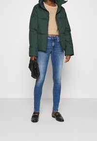 Chaqueta de plumas verde con cuello alto, suéter de punto beige, jeans ajustados azules y mocasines negros. Bolso negro en la mano.