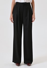 Zwarte, hooggewaiste broek met een wijde pijpdesign en plooien aan de voorkant. Gemaakt van een gladde stof, gecombineerd met puntige rode schoenen.