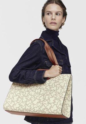 Frau in dunkler Jeansjacke, die eine große beige Tragetasche mit abstraktem braunem Muster und breiten braunen Trägern über der Schulter trägt.