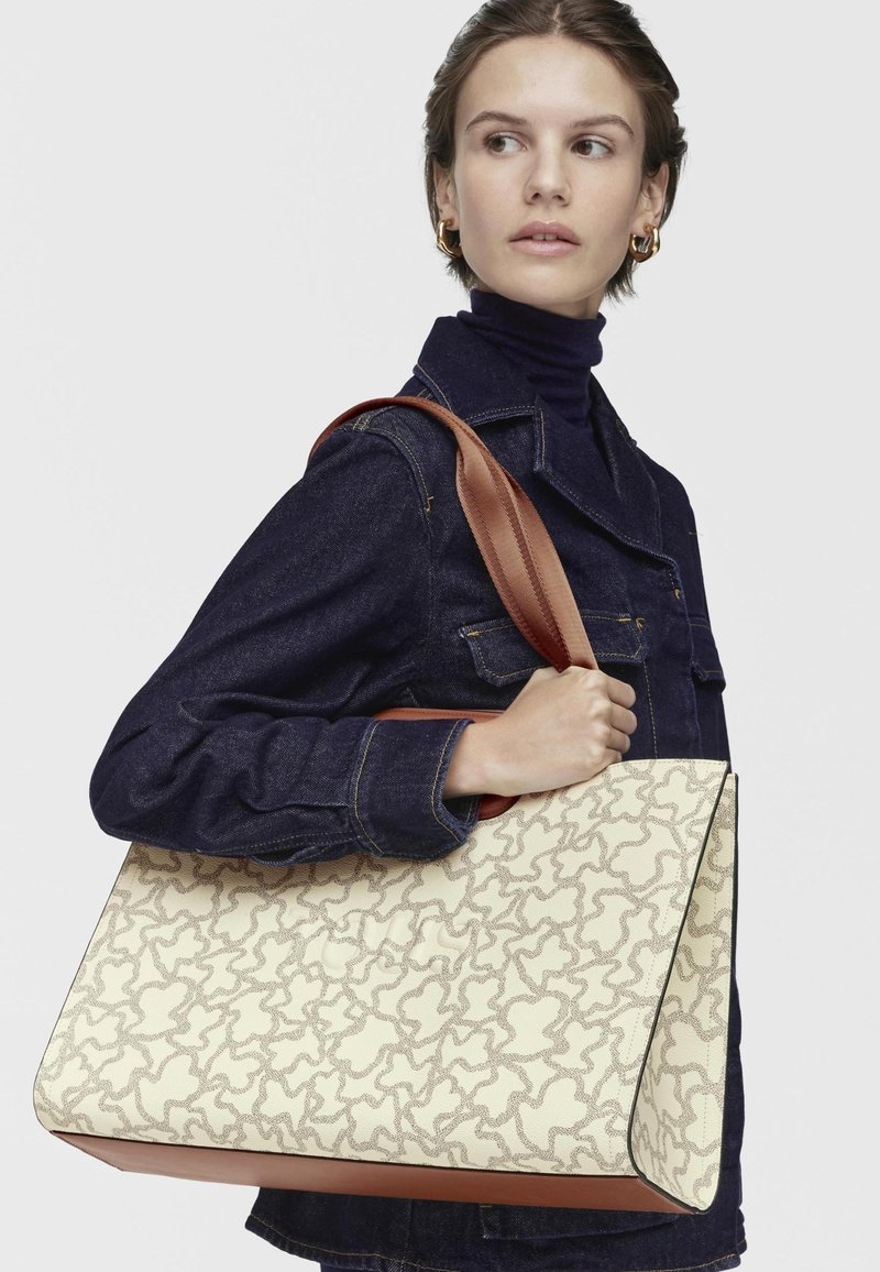 Mujer con chaqueta de mezclilla oscura llevando un gran bolso beige con un patrón abstracto en marrón y asas anchas marrones sobre el hombro.