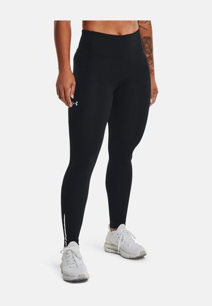Persoon draagt zwarte leggings met hoge taille en een zijlogo van Under Armour, en witte sportschoenen, staand tegen een effen achtergrond.