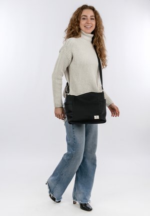 Sac à épaule noir avec texture lisse, deux poches latérales et détail de logo. L'utilisateur porte un pull léger et un jean évasé avec des bords effilochés.