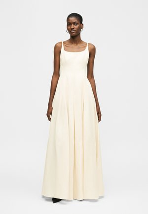 Μαύρη γυναίκα που φοράει αμάνικο, μακρύ μέχρι το πάτωμα φόρεμα σε κρεμ απόχρωση με τετράγωνη λαιμόκοψη, στέκεται μπροστά σε απλό φωτεινό φόντο, κοιτάζοντας προς τα εμπρός.