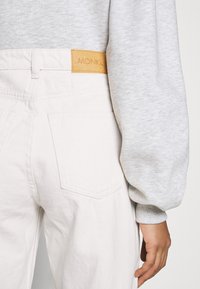 Jeans de ganga branca com um bolso traseiro visível e uma etiqueta de couro bege marcada com "MONKI". Um fato de treino cinzento está parcialmente visível.