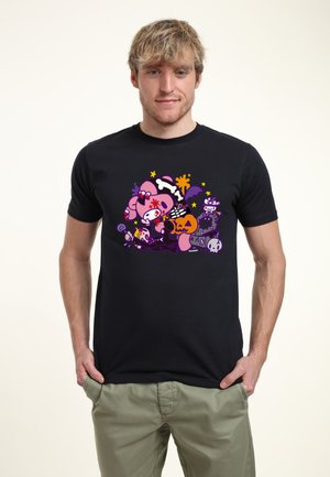 Jeune homme portant un T-shirt noir avec un design coloré sur le thème d'Halloween incluant des citrouilles, des crânes et des fantômes, debout les mains dans les poches.
