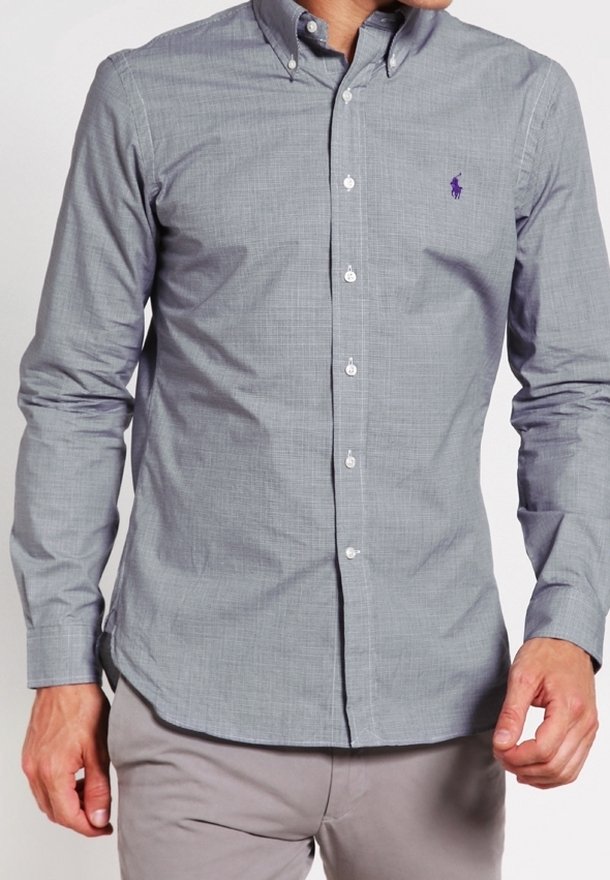 Man i ljusgrå skjorta med knappar och en lila häst-logotyp på bröstet, samt ljusgrå byxor, står med avslappnade armar.