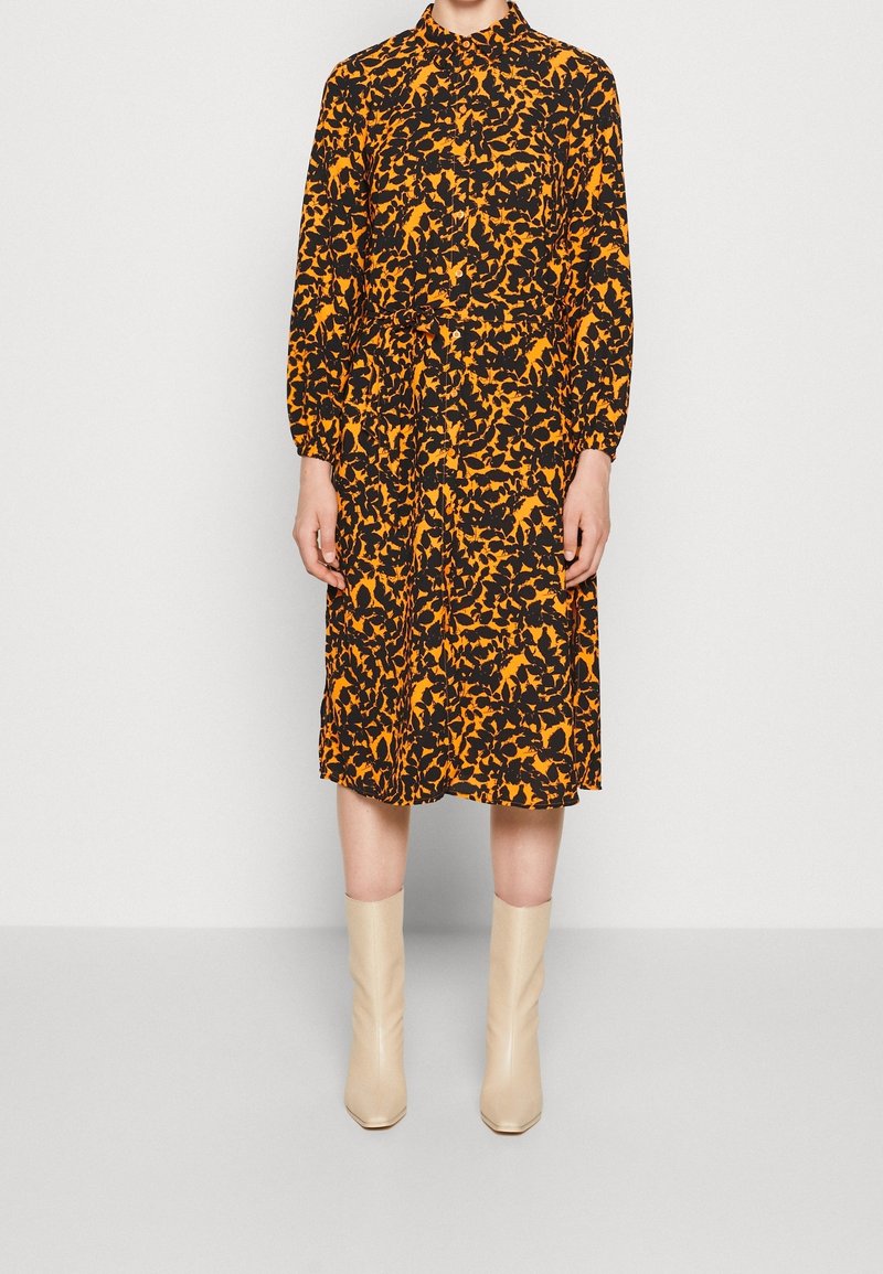 Robe noire avec un motif abstrait orange, manches longues, col et ceinture à la taille, assortie à des bottines crème à talons hauts.