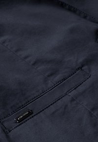 Marinblå jacktyg med slät textur, med en horisontell ficka och en liten svart metallmärkning sydd framtill.