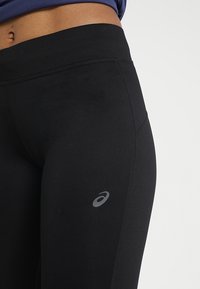 Czarne legginsy sportowe wykonane z elastycznego materiału; mają gładką teksturę i subtelne logo na dolnym lewym boku; wysoki fason passa.