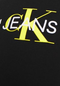 Černá tkanina s výrazným textem "JEANS" v bílé barvě a velkým "K" v neonově žluté, s moderním a grafickým designem.