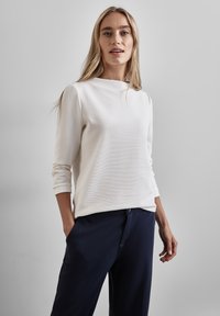 Top blanco de manga larga con un cuello alto, textura sutil y puños ajustados, combinado con pantalones azul marino. Diseño limpio, aspecto moderno.