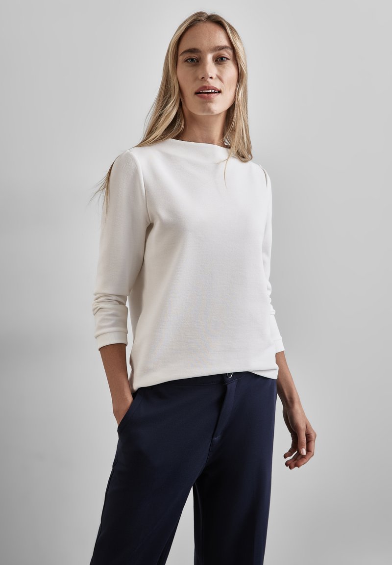 Top blanco de manga larga con un cuello alto, textura sutil y puños ajustados, combinado con pantalones azul marino. Diseño limpio, aspecto moderno.