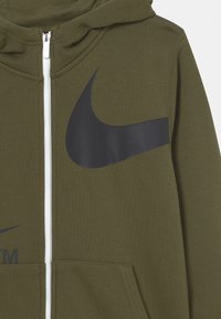 Olivově zelená mikina na zip z měkké látky. Má velké černé logo Nike Swoosh na hrudi a přední kapsy. Bílý zip.