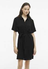 Juoda suknelė marškiniai su apykakle, trumpomis rankovėmis, mygtukais priekyje, surišta juosmeniu ir šiek tiek tekstūruota medžiaga.