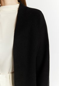 Zwarte cardigan van zacht materiaal, met een ontspannen pasvorm, open voorkant en naadloze schouders, gecombineerd met een crèmekleurige blouse eronder.