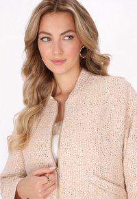 Roze textuurjasje met metalen draad, ritssluiting aan de voorkant en zijzakken. Model draagt het over een lichte top met minimale accessoires.