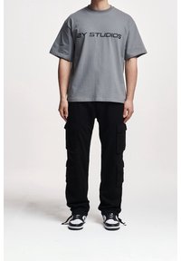 Harmaa puuvillainen t-paita, jossa on mustat kirjaimet "2Y STUDIOS", yhdistettynä mustiin cargo-housuihin, joissa on useita sivutaskuja, ja nyörillisiin tennareihin.