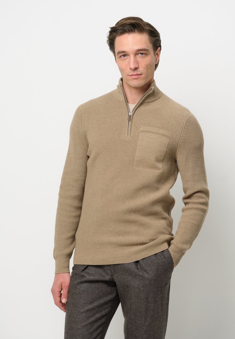 Beige gestrickter Pullover mit einem halben Reißverschluss und Brusttasche. Rippenstruktur, taillierte Passform, kombiniert mit grauen Hosen.