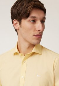 Camisa amarilla de botones, hecha de una tela suave, con un logo bordado de un dachshund blanco en el bolsillo izquierdo del pecho.