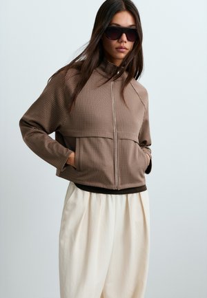 Femme portant de grandes lunettes de soleil foncées, une veste marron à fermeture éclair avec un motif texturé, et un pantalon blanc ample, debout les mains dans les poches.