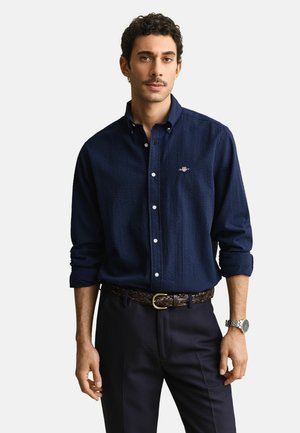 Mann trägt ein dunkelblaues, strukturiertes Langarmhemd mit hochgekrempelten Ärmeln, dunkelblaue Hose, geflochtener Gürtel und silberne Armbanduhr.