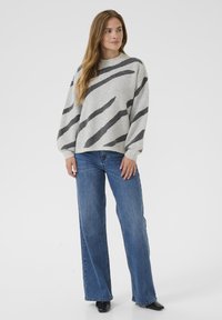 Femme debout portant un pull gris clair avec des rayures diagonales noires, un jean large bleu et des chaussures pointues noires, sur un fond uni.