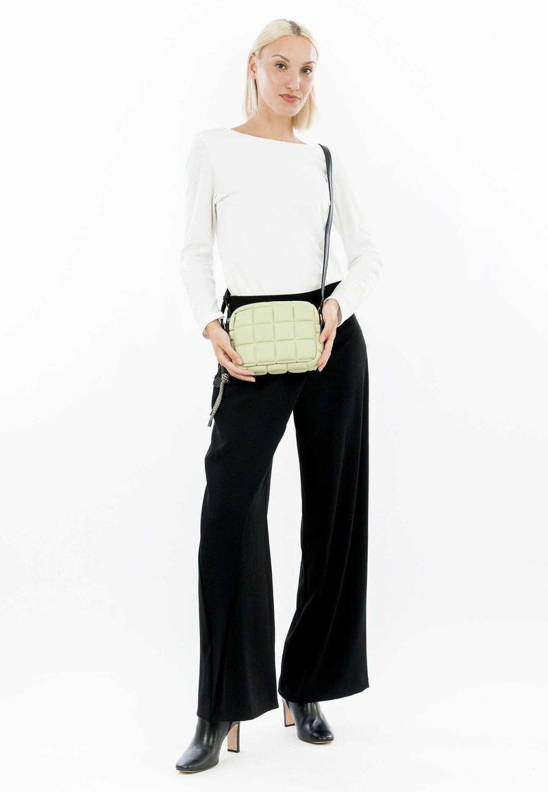 Bolso cruzado acolchado verde con un diseño de acolchado; cuenta con una correa ajustable negra y cierre de cremallera. Modelo con pantalones negros y blusa blanca.