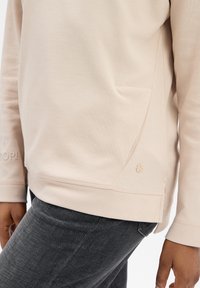 Sudadera beige de manga larga, con textura suave, bolsillo lateral y un sutil detalle de logo. Combinada con jeans gris oscuro.