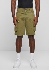 Pantaloni scurți cargo verde măsliniu, cu buzunare laterale, închidere cu nasturi și bretele, fabricați dintr-un material robust. Purtați cu adidași albi și șosete.