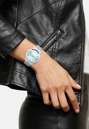 Sølv ur med en rund skive, mintfarvet urskive, sølv timeindikatorer og et armbånd i rustfrit stål, båret på en hånd mod en sort læderjakke.