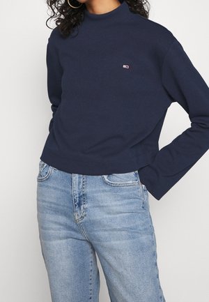 Frau in marineblauem Langarmshirt mit kleinem Logo auf der Brust und hellblauer hochtaillierter Jeans, die mit einer Hand in der Tasche steht.