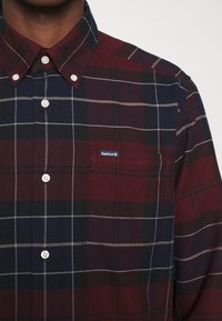 Homme portant une chemise à carreaux rouge foncé et bleu marine boutonnée, avec une poche avant étiquetée « Barbour ».