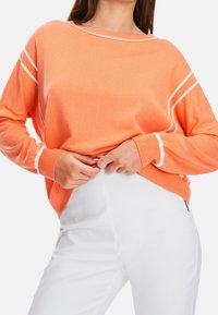 Pull orange léger avec accents blancs, large encolure et manches longues ; associé à un pantalon blanc ajusté. Texture en maille douce.