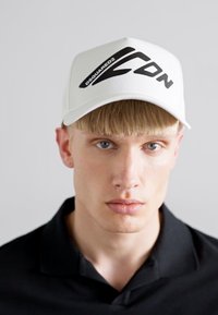 Cappello bianco con logo e dettagli neri "D-Squared2". Tessuto morbido, visiera curva, forma strutturata, indossato con una polo nera.
