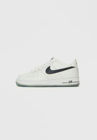 Nike Air Force 1 tossud, millel on valgest nahast pealis, must Swooshi logo, ümmargune varvas ja läbipaistev kummist välistald.