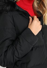 Giacca imbottita nera con chiusura a zip e cappuccio con pelliccia sintetica. Presenta sezioni trapuntate e una texture morbida e liscia. Dolcevita rossa sotto.