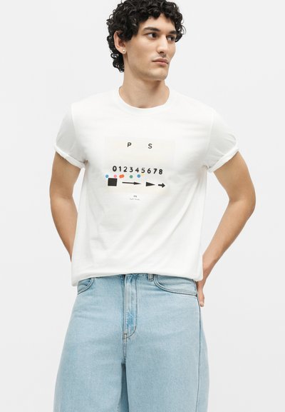 Άνδρας με σγουρά μαλλιά που φοράει λευκό T-shirt με γραφικά αριθμών και πολύχρωμες κουκκίδες, σε συνδυασμό με ανοιχτόχρωμο τζιν παντελόνι.