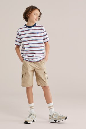 Niño de pie con las manos en los bolsillos, vestido con camiseta de rayas, pantalones cortos cargo beige, calcetines blancos con rayas marrones y zapatillas beige.