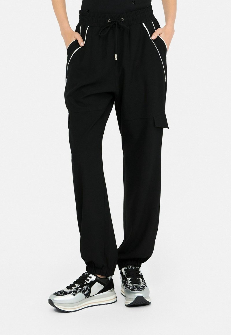 Pantalon de jogging noir avec passepoil blanc, taille et poignets élastiques, poche cargo latérale ; porté avec des baskets à plateforme noires et argentées.