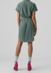 Vihreä, lyhythihainen mekko, jossa on vyötärösolki ja pyöristetty helma. Yhdistetty vaaleanpunaisten korokepohjakenkien ja vaaleansinisillä resorisukilla.