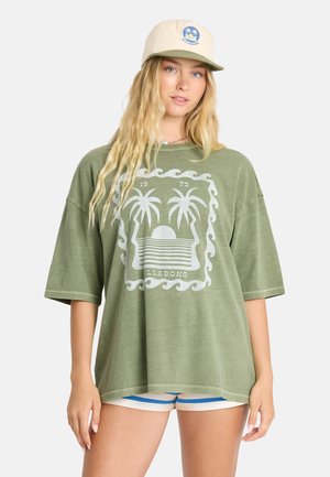 Jonge vrouw met lang blond haar die een groene oversized t-shirt met palmboomprint, een beige pet en blauw-witte korte broek draagt.