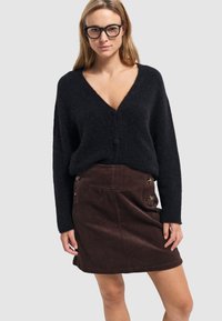 Zwarte, oversized v-hals cardigan van zachte stof, gecombineerd met een bruine ribfluwelen rok met knoopaccenten aan de voorkant en een gestructureerd oppervlak.