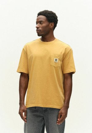 Homme aux cheveux courts et bouclés portant un t-shirt jaune moutarde avec une poche poitrine et un jean gris, regardant vers sa gauche.