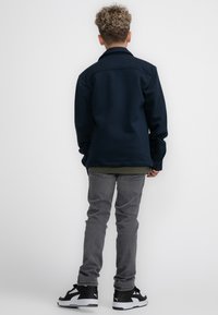 Marine oversized jasje met een kraag, grijze jeans en zwarte sneakers met witte accenten. Kruidachtig haar, gezien vanuit de rug.